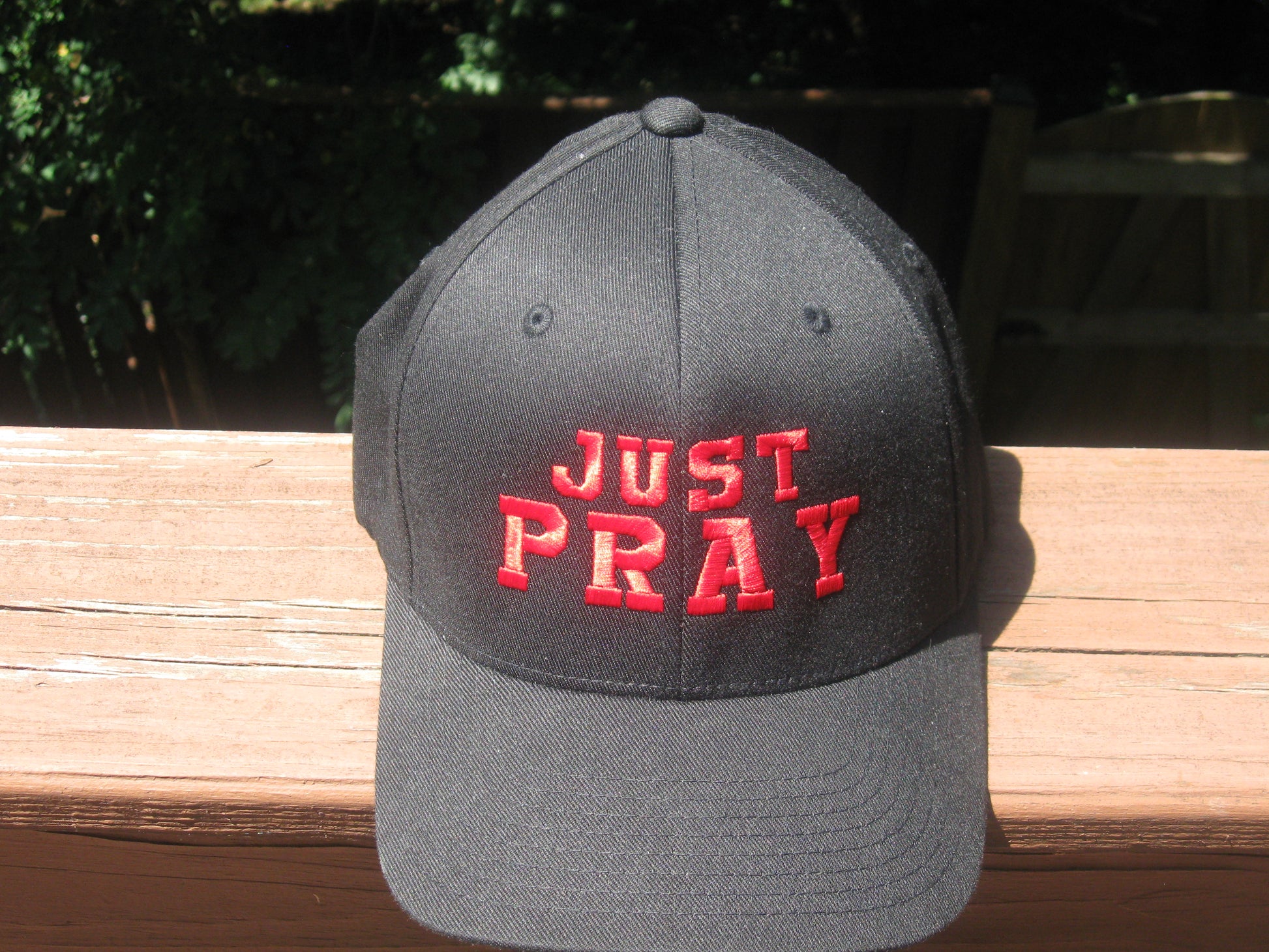 A structured, six-panel twill cap in a black featuring the words "Just Pray" embroidered on the front. The cap has a low-profile embroidery area, six vent eyelets for breathability. It is displayed on a neutral background.